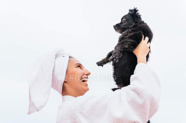Оптимістична молода жінка в халаті і рушнику посміхається і приймає чорного собаку під час спа-сеансу вдома — стокове фото