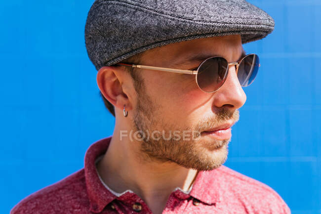 Молодой бородатый парень в обычной красной рубашке поло и кепке, стоящий напротив синей стены на улице в летний день — стоковое фото