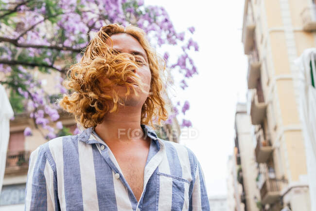 From below of dreamy curious male with long wavy hair standing in street in summer with flower tree — Stock Photo