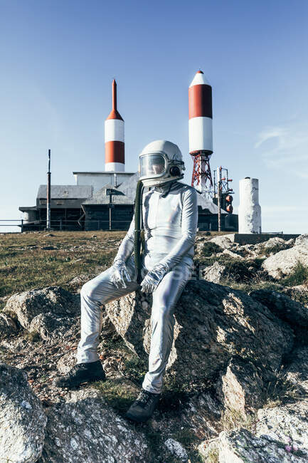 Hombre de cuerpo completo en traje espacial sentado en el suelo rocoso rayas cohete en forma de antenas en el día soleado - foto de stock