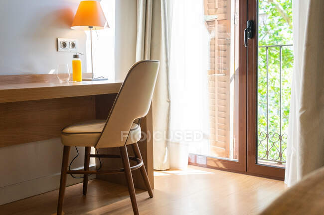 Comfortable chair placed near table with glass bottle of fresh juice in room with curtains hanging on glass doors to balcony — Stock Photo