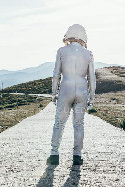 Hombre de vista trasera en traje espacial y casco mirando hacia otro lado mientras está parado en el camino en el día soleado en la naturaleza - foto de stock