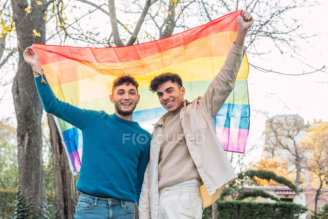 Низкий угол веселой парочки гомосексуальных мужчин, стоящих с радужным ЛГБТ-флагом, машущим над головами в парке и смотрящих друг на друга — стоковое фото