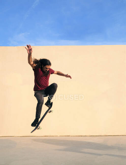 Чоловік скейтбордист з хвилястим волоссям виконує трюк на скейтборді, стрибаючи через прохід і дивлячись вниз в сонячний день з блакитним небом — стокове фото