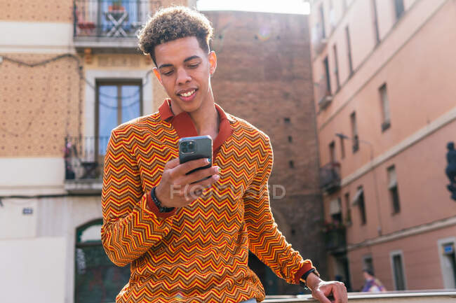 Von unten ein junger hispanischer Typ mit Afro-Haaren in stylischem, buntem Outfit, der im Sonnenlicht auf einem Geländer in der Nähe eines städtischen Gebäudes steht und sein Handy surft — Stockfoto