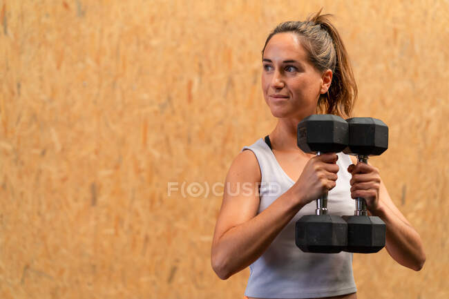 Focused slim female athlete doing exercise with heavy dumbbells during training in gym — Stock Photo