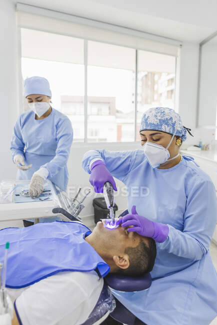 Doctor wearing medical uniform treating client with dental tool with assistant preparing instruments in hospital — Stock Photo