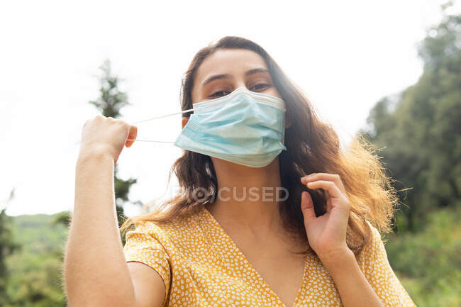Низкий угол улыбки женщины надевает защитную медицинскую маску во время коронавируса и смотрит на камеру в природе летом — стоковое фото