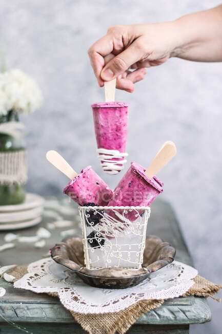 Mulher sem rosto em vestido branco organizando deliciosos picolés de iogurte de amora-preta caseiros em cesta com gelo na mesa rústica — Fotografia de Stock