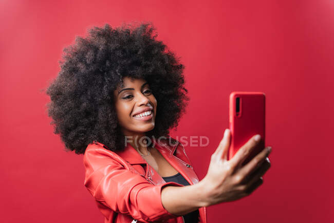 Lächelnde Afroamerikanerin mit Afro-Frisur beim Selbstporträt auf Handy auf rotem Hintergrund im Studio — Stockfoto
