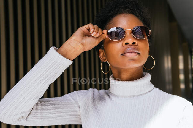 Молода афроамериканська жінка в стильному светрі і сонцезахисних окулярах, дивлячись у далечінь, стоячи на яскравому світлі проти металевого тла. — стокове фото