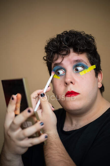 Concentrado excêntrico feminino masculino aplicando cor olhos com escova ao fazer maquiagem e segurando espelho — Fotografia de Stock