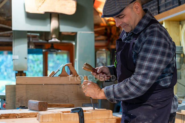 Side view woodworker with wooden hammer and chisel carving wooden detail while working in professional joinery workshop — Stock Photo