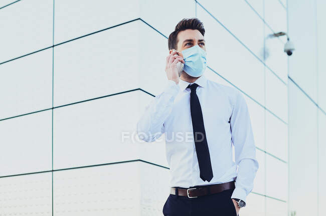 Elegante ejecutivo masculino en ropa formal y máscara médica hablando por teléfono celular mientras mira hacia otro lado en la ciudad - foto de stock