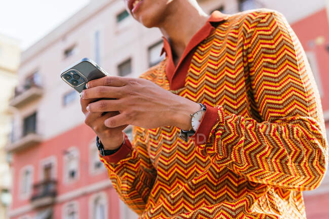 From below cropped unrecognizable side view of young Hispanic guy with Afro hair in stylish colorful outfit browsing mobile phone while standing on railing near urban building in sunlight — Stock Photo