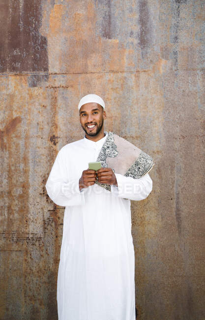 Allegro maschio musulmano in abiti tradizionali sorridente e navigando cellulare mentre in piedi vicino al muro squallido sulla strada — Foto stock