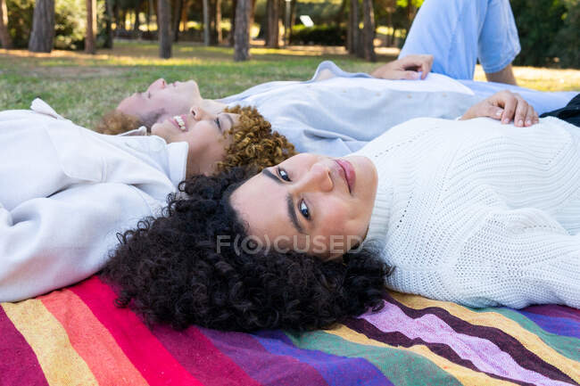 Diverse women and man with curly hair lying face to face on colorful plaid in park looking at camera — Stock Photo