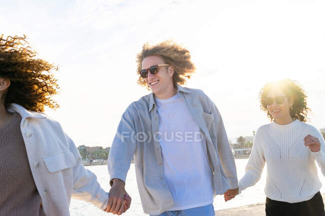 Grupo de mujeres jóvenes y diversos hombres con el pelo rizado tomados de la mano mientras caminan en la costa del paisaje urbano en la espalda iluminada - foto de stock
