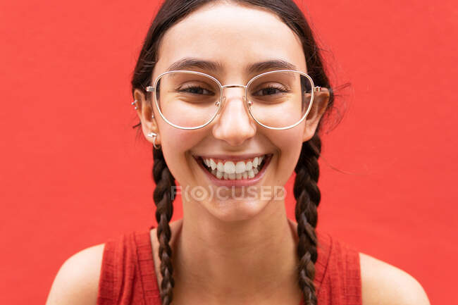 Fröhliche junge Frau mit Zöpfen Frisur, während sie in die Kamera auf rotem Hintergrund in der Straße schaut — Stockfoto