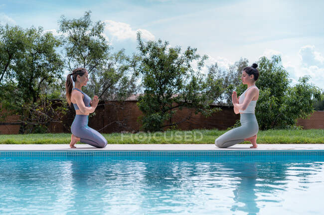 Vista lateral de las mujeres en ropa deportiva realizando pose en cuclillas de los pies mientras practican yoga con las manos orantes entre la piscina y el césped - foto de stock