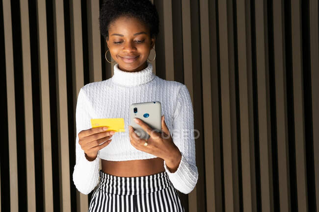 Positive junge Afroamerikanerin in trendiger Kleidung lächelt und bezahlt mit Kreditkarte und Smartphone für Online-Bestellungen gegen die gestreifte Wand auf der Straße — Stockfoto