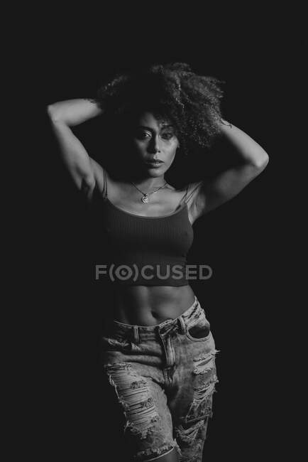 Curly haired African American female in trendy crop top and jeans standing touching hair on black background in studio — Stock Photo