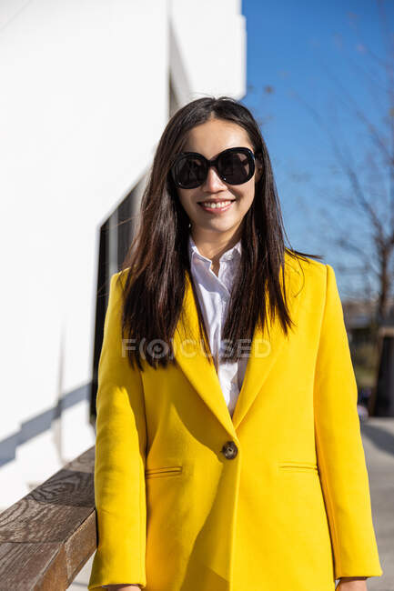 Sorridente donna d'affari asiatica con cappotto giallo che cammina sulla strada con edificio sullo sfondo — Foto stock