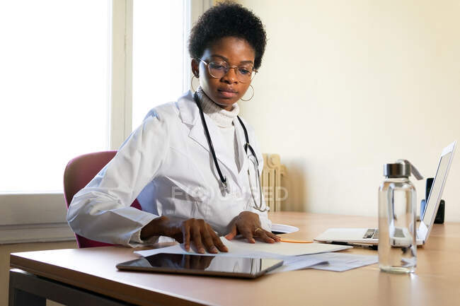 Joven médica afroamericana en bata blanca con estetoscopio sentada en la mesa con portátil y leyendo registros médicos mientras trabaja en la clínica - foto de stock