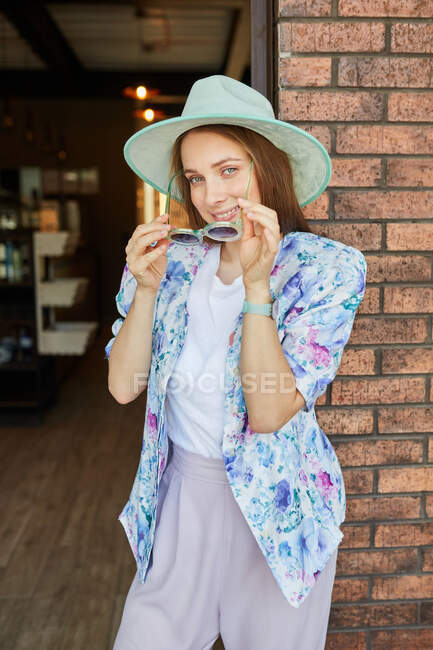 Joyeux jeune femme en tenue tendance avec des lunettes de soleil regardant la caméra contre un mur rugueux en ville — Photo de stock