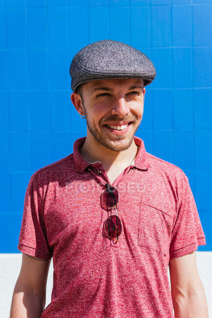 Радостный молодой бородатый мужчина в стильной повседневной одежде и кепке наслаждается летним днем возле голубой стены на городской улице — стоковое фото