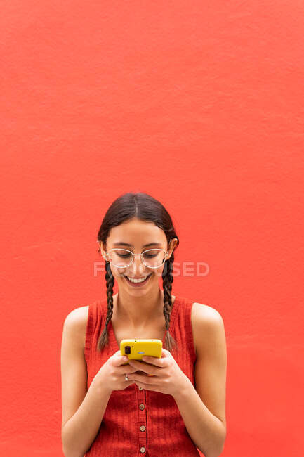 Cheerful young female in pigtails hairstyle browsing on smartphone standing on red background in street — Stock Photo