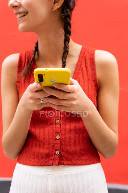 Schnitte unkenntlich anonyme junge Frau mit Zöpfen Frisur suren auf dem Smartphone stehen wegschauen auf rotem Hintergrund in der Straße — Stockfoto
