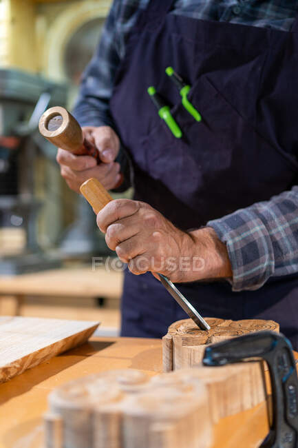 Unrecognizable male woodworker with wooden hammer and chisel carving wooden detail while working in professional joinery workshop — Stock Photo
