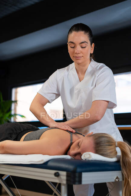 Masseuse amicale souriant et massant les épaules de la femme tout en travaillant dans une clinique de physiothérapie — Photo de stock
