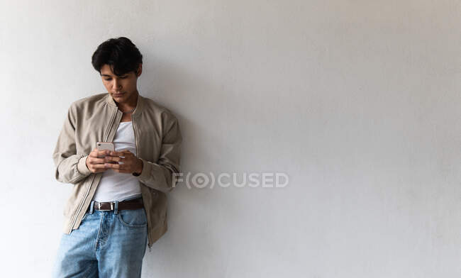 Giovane focalizzato uomo latino in abiti casual utilizzando il telefono mentre appoggiato sulla parete bianca — Foto stock