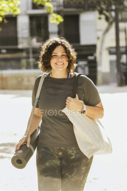 Позитивний молодий плюс розмір жінки з кучерявим темним волоссям в повсякденному одязі посміхається під час прогулянки по міській вулиці з прокатом мат перед сеансом йоги в сонячний ранок — стокове фото