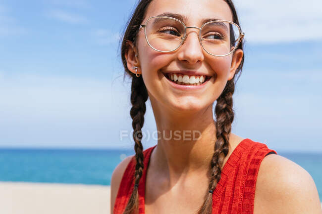 Cheerful female in summer clothes with pigtails standing on sandy shore with calm blue sea on sunny day — Stock Photo