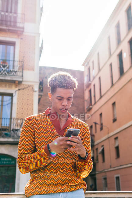 Giovane ragazzo ispanico con i capelli afro in elegante vestito colorato navigazione cellulare mentre in piedi su ringhiera vicino edificio urbano alla luce del sole — Foto stock