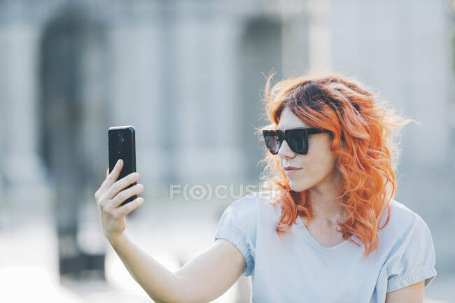 Donna alla moda con i capelli rossicci e in occhiali da sole prendendo auto colpo sul telefono cellulare nella giornata di sole in strada — Foto stock
