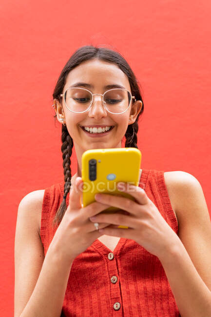 Alegre joven hembra en coletas peinado navegación en el teléfono inteligente de pie sobre fondo rojo en la calle - foto de stock