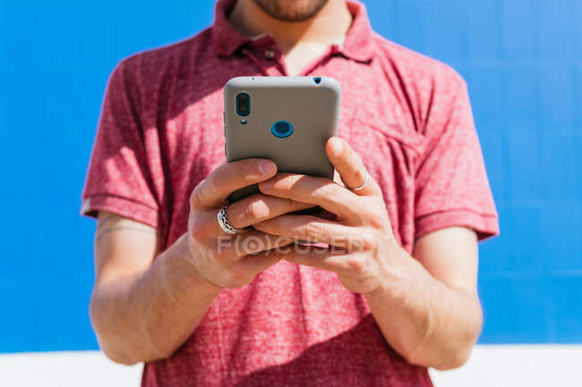 Обрезанный неузнаваемый хипстер в повседневной рубашке для поло и кепке, просматривающий мобильный телефон, стоя напротив синей стены в солнечном свете, отворачиваясь — стоковое фото