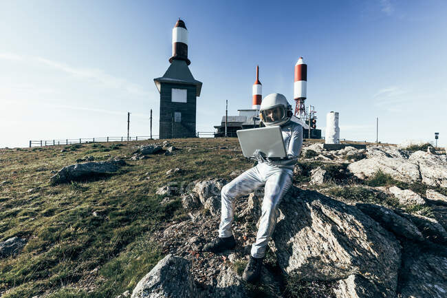 Astronauta masculino de cuerpo completo en traje espacial navegando datos en netbook mientras está sentado en la roca fuera de la estación con antenas en forma de cohete - foto de stock