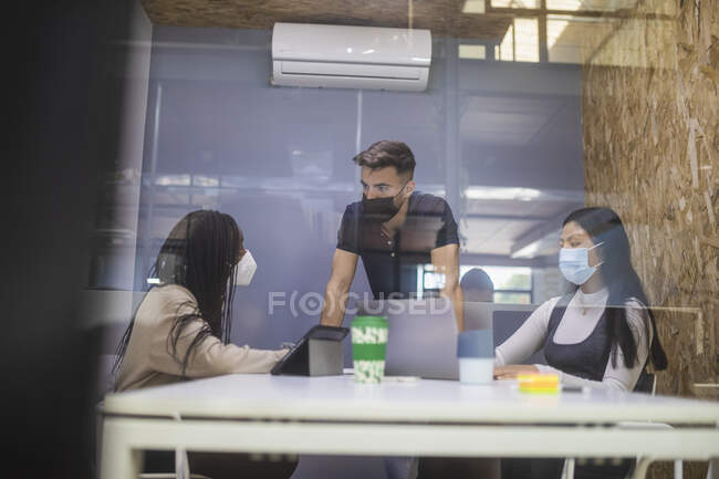 Through glass of company of multiracial coworkers in protective masks gathering at table and discussing project during business meeting — Stock Photo