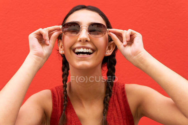 Jovem encantada com tranças colocando óculos de sol na moda contra o fundo vermelho na rua da cidade — Fotografia de Stock