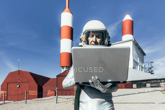 Чоловічий астронавт у скафандрі переглядає дані з нетбука, стоячи надворі з антеною, що має форму ракети. — стокове фото