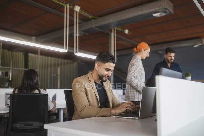 Alegre administrador masculino navegar netbook mientras está sentado en la mesa y trabajar en el espacio de coworking con colegas irreconocibles - foto de stock