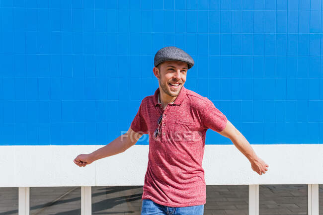 Gioioso giovane maschio barbuto in elegante abbigliamento casual e cappello divertirsi e godersi la giornata estiva vicino al muro blu sulla strada urbana — Foto stock
