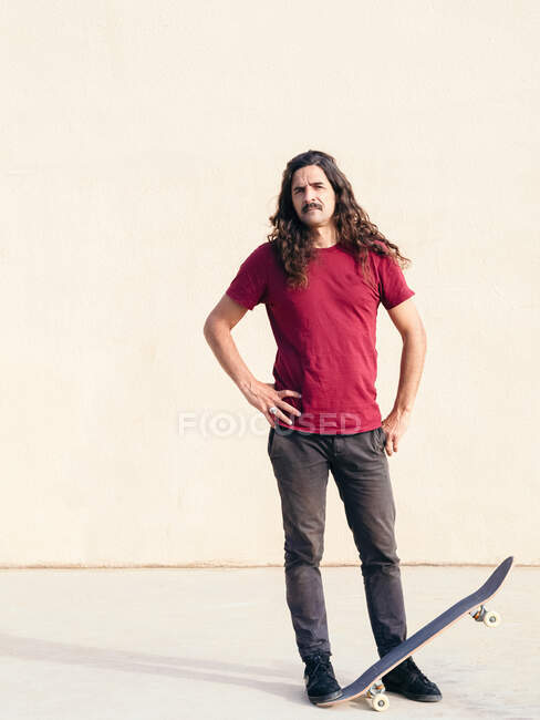Чоловічий скейтбордист з вусами і довгим волоссям з руками на стегнах стоїть, дивлячись на камеру на тротуарі на сонячному світлі на бежевому фоні — стокове фото