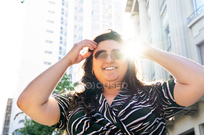 Low angle of confident young curvy female in stylish dress with geometric print and trendy sunglasses looking at camera while standing near stone urban building in summer day — Stock Photo