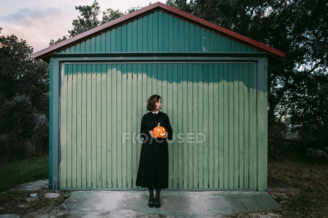 Female in black dress and with Halloween pumpkin lantern standing against wooden wall of house and looking away — Stock Photo